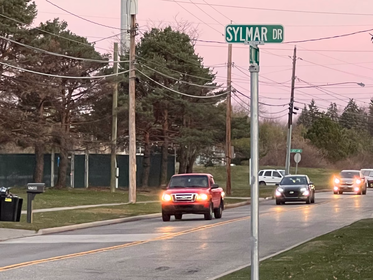 Cars driving on Mill Road and a street sign for Sylmar Drive, which connects houses to the road.